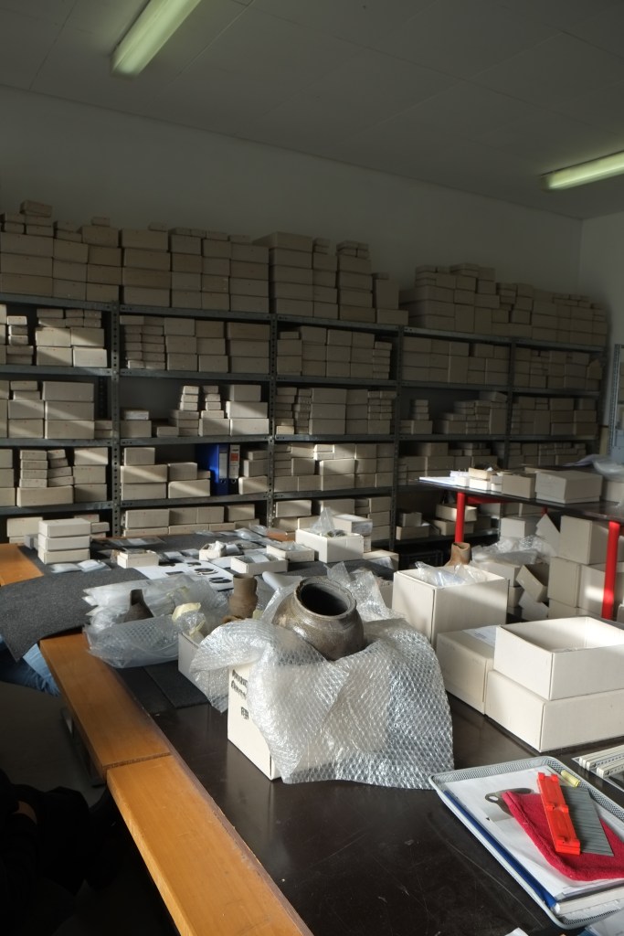 Auf einem großen Tisch stehen geschlossene und geöffnete Kisten, aus denen zum Teil Funde zu sehen sind. Im Hintergrund ein großes Regal mit Kartons unterschiedlicher Größen.