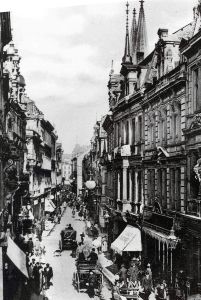 Historische schwarz-weiß Aufnahme mit Kutschen auf der Straße und den historischen Gebäuden. Rechts hinter den Häuserfassaden sind die Domspitzen zu sehen.