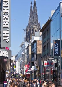 Foto der gut besuchten Einkaufsstraße mit Geschäftsschildern und rechts im Hintergrund die Spitzen des Kölner Doms.