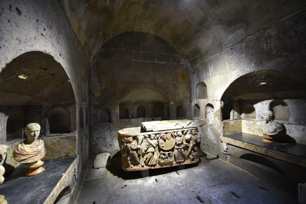 Gewölbe mit einem steinernen Sarkophag in der Bildmitte. Rechts und links römische Büsten.