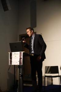 Vortragender am Stehpult vor einem Laptop. Er spricht in ein Mikrofon.