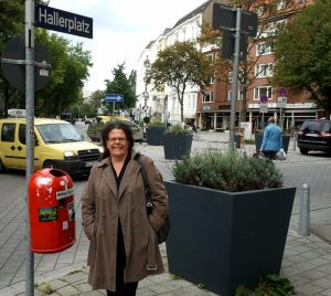 Frontalaufnahme der lächelnden Annette Haller an einem Straßenschild mit der Aufschrift "Hallerplatz".