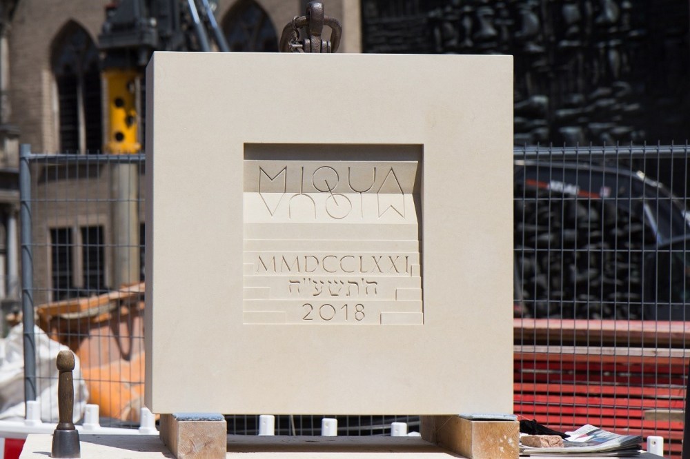 Der MIQUA-Grundstein aus Kalkstein in Form eines Quadrates. In der Mitte des Steins hervorgehoben der gespiegelte Schriftzug MIQUA sowie über Stufen dargestellt verschiedene Zeitrechnungen.