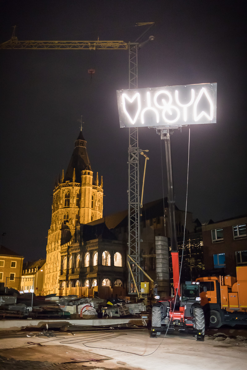 Das Logo des MiQua wurde mithilfe eines Krans nach oben gezogen und leuchtet über der Baustelle. Im Hintergrund sind die Renaissance-Laube und der Rathaus Turm zu sehen.