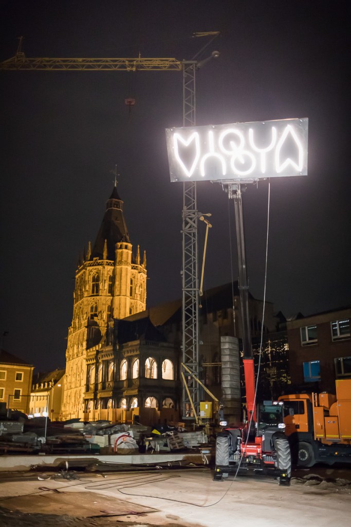 Das Logo des MiQua wurde mithilfe eines Krans nach oben gezogen und leuchtet über der Baustelle. Im Hintergrund sind die Renaissance-Laube und der Rathaus Turm zu sehen.