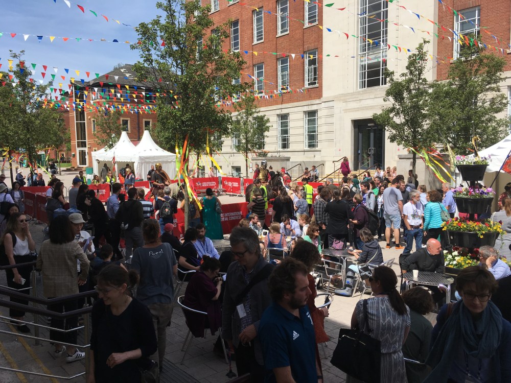 Der freie Platz zwischen den Häuserreihen ist mit Menschen gefüllt. In der Mitte sind Sitzgelegenheiten aufgebaut und ein Bereich für Reiter in mittelalterlicher Kleidung auf ihren Pferden eingegrenzt. Girlanden reichen von Haus zu Haus.