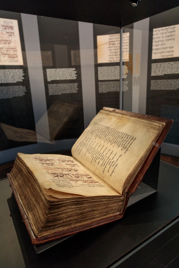 Der Amsterdam Machsor ist eine hebräische Handschrift aus dem Mittelalter. Das Foto zeigt das Manuskript aufgeschlagen in der Vitrine liegend. Im Hintergrund sind Teile der Ausstellungsarchitektur erkennbar.