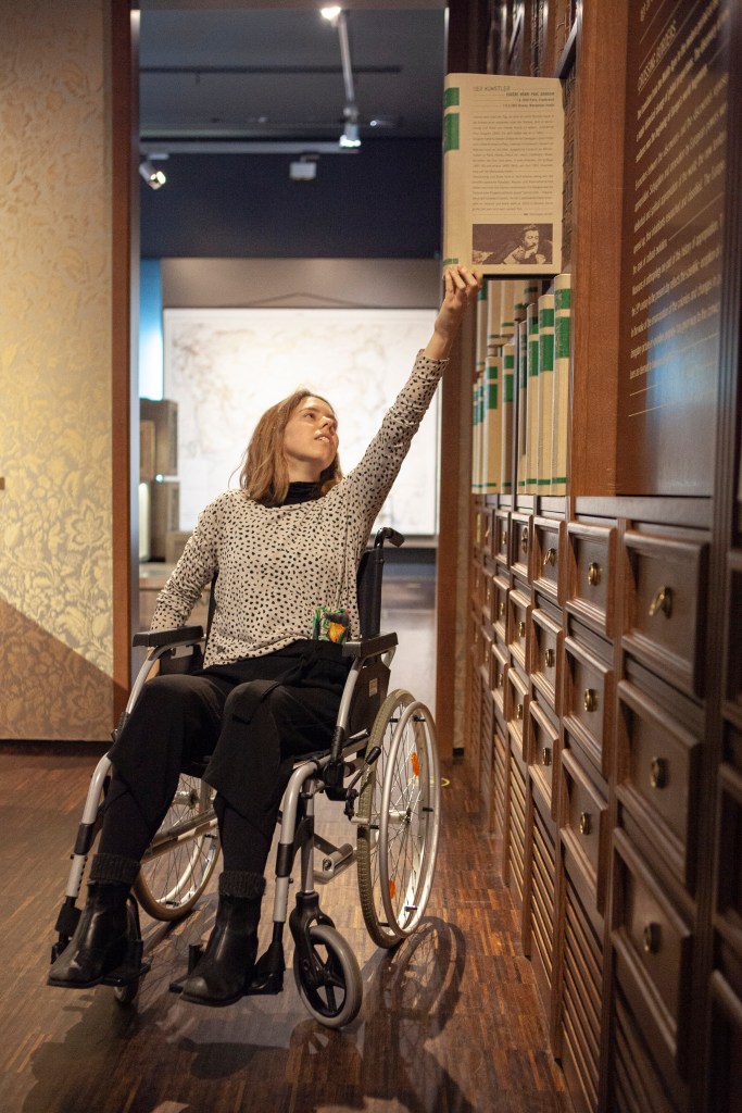 Eine MiQua Kollegin versucht, im Rollstuhl sitzend, interaktive Elemente in der Ausstellung zu nutzen. Dabei muss sie sich ganz hochrecken und seitlich an die Bücherwand mit ausziehbaren Elementen heranfahren.