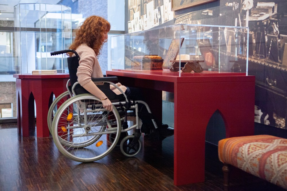 Eine Kollegin unterfährt mit dem Rollstuhl eine Vitrine. Sie kann die Exponate nur aus der Nähe sehen, weil der Sockel der Vitrine nicht geschlossen ist.