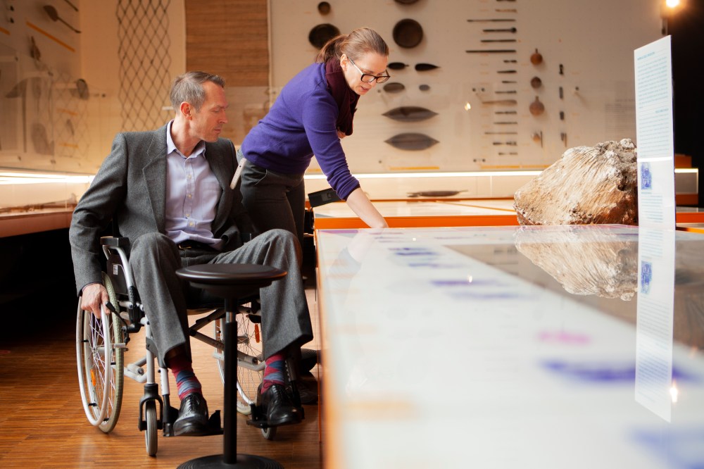 MiQua Direktor Thomas Otten testet im Rollstuhl sitzend einen Multimedia-Tisch in der Ausstellung. Wie unterschiedlich die Perspektive ist, wird über eine Kollegin deutlich, die neben ihm steht.