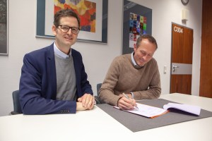 Die Direktoren von APG und MiQua, Marco Lohmann und Thomas Otten, sitzen an einem weißen Tisch und sehen die Kooperationsvereinbarung durch. Beide lächeln und schauen seitlich in Richtung des Fotografen. Im Hintergrund hängen Gemälde an der Wand des Büros.