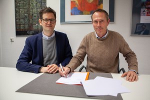 Der Direktor des APG Marco Lohmann, links und Thomas Otten, Direktor des MiQua rechts im Bild, sitzen an einem weißen Tisch und unterzeichnen die Kooperationsvereinbarung. Beide schauen gut gelaunt in die Kamera. Im Hintergrund hängen Gemälde an der Wand des Büros.