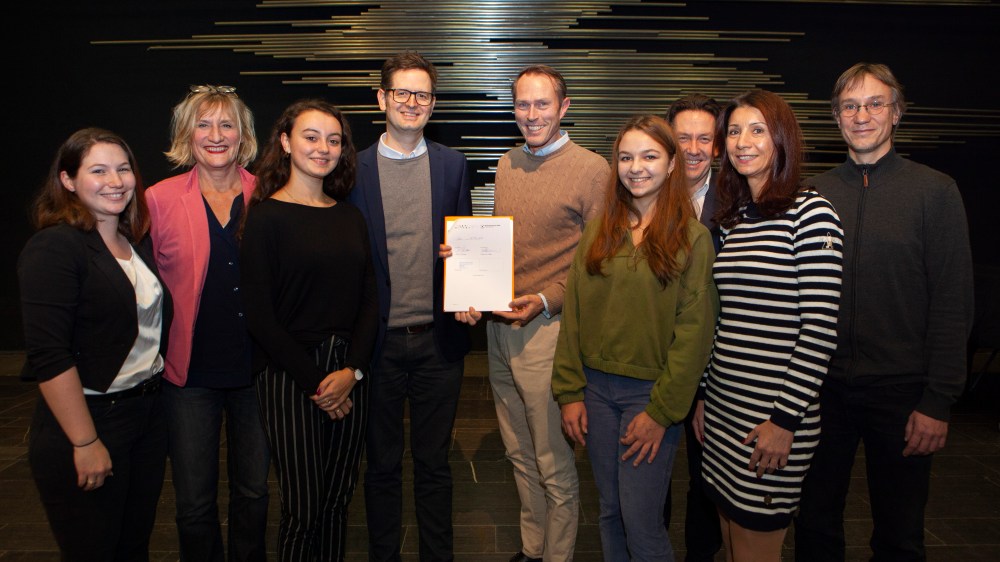 Insgesamt neun Personen - fünf Frauen, vier Männer - waren bei der Unterzeichnung dabei und präsentieren auf diesem Bild die unterzeichnete Kooperationsvereinbarung. Sie alle stehen auf der Bühne des APG. Im Hintergrund ist das Relief "Eine Flächenbahn" von Norbert Kricke zu sehen.