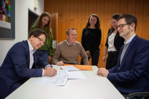 Frank Schweppenstette, links im Bild, sitzt am Konferenztisch und unterschreibt die Kooperationsvereinbarung zur Bildungspartnerschaft. Ebenfalls dabei sind die anderen Unterzeichner*innen. Sie alle sind um den Tisch versammelt und gut gelaunt.
