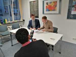 Im Vordergrund des Bildes positioniert sich der Fotograf, um die beiden Direktoren bei der Unterzeichnung abzulichten. Die beiden Herren sitzen im Bildzentrum an einem Tisch und sehen das zu unterzeichnende Papier durch. Der Fotograf ist nur von hinten zu sehen.