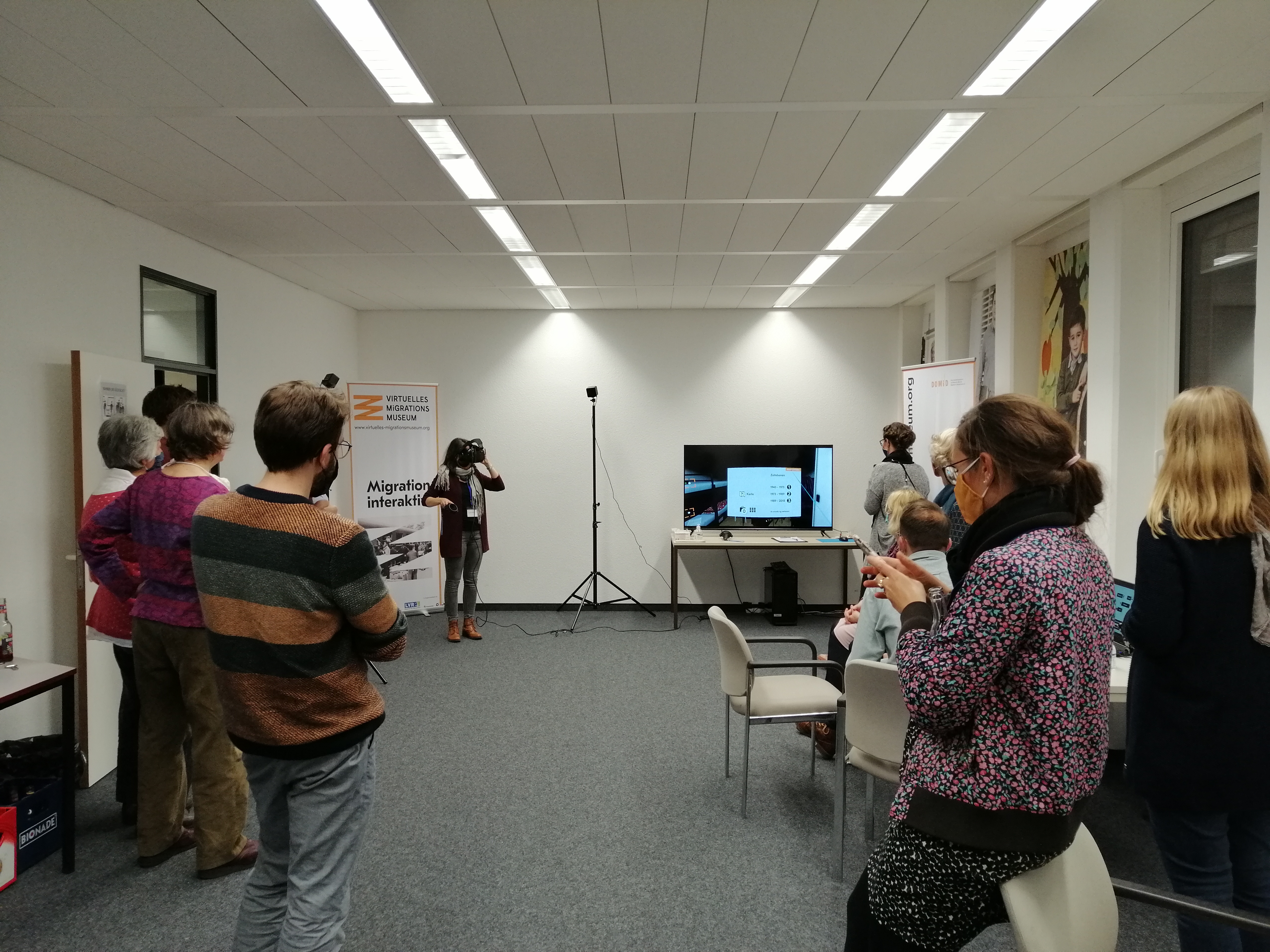 Blick in den Veranstaltungsraum des DOMiD. Am linken und rechten Bildrand verteilt stehen die Teilnehmer*innen und folgen der Präsentation von Sandra Vacca. Sie - hinten links im Bild mit VR-Brille und Steuerung - zeigt über einen Bildschirm wie die Anwendung als Virtual Reality-Angebot funktioniert.