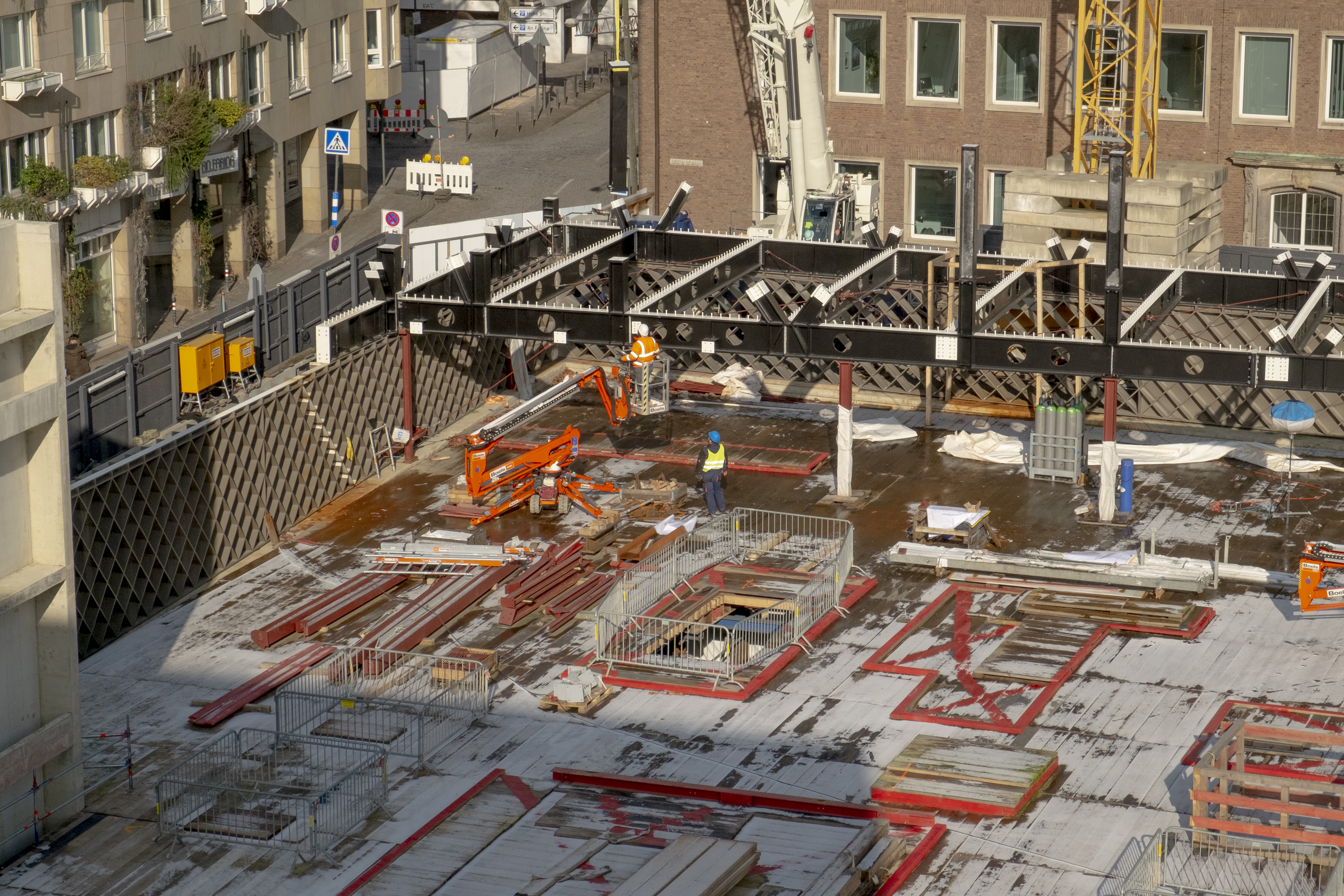 Die MiQua Baustelle ist Anfang Februar mit einer dünnen Schnee- oder Eisschicht überzogen. Der Blick von Süden auf den Baustellenabschnitt an der Portalsgasse und Unter Goldschmied zeigt, dass der Grundriss des späteren Gebäudes schon mit den sogenannten Rautentragwerken, einer Stahlkonstruktion, angezeigt wird. Die Arbeiter auf der Baustelle arbeiten in diesem Moment an einem Querträger, der später das Obergeschoss halten wird. Den oberen Abschluss des Bildes, bildet der Spanische Bau. Links im Bild ist noch einer der Aufzugschächte aus Beton erkennbar.