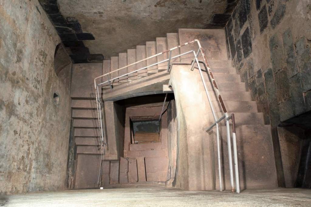 Blick in einen viereckigen Raum, an dessen Wänden eine Treppen in die Tiefe führt. Am Boden erkennt man ein viereckiges mit Wasser gefülltem Becken