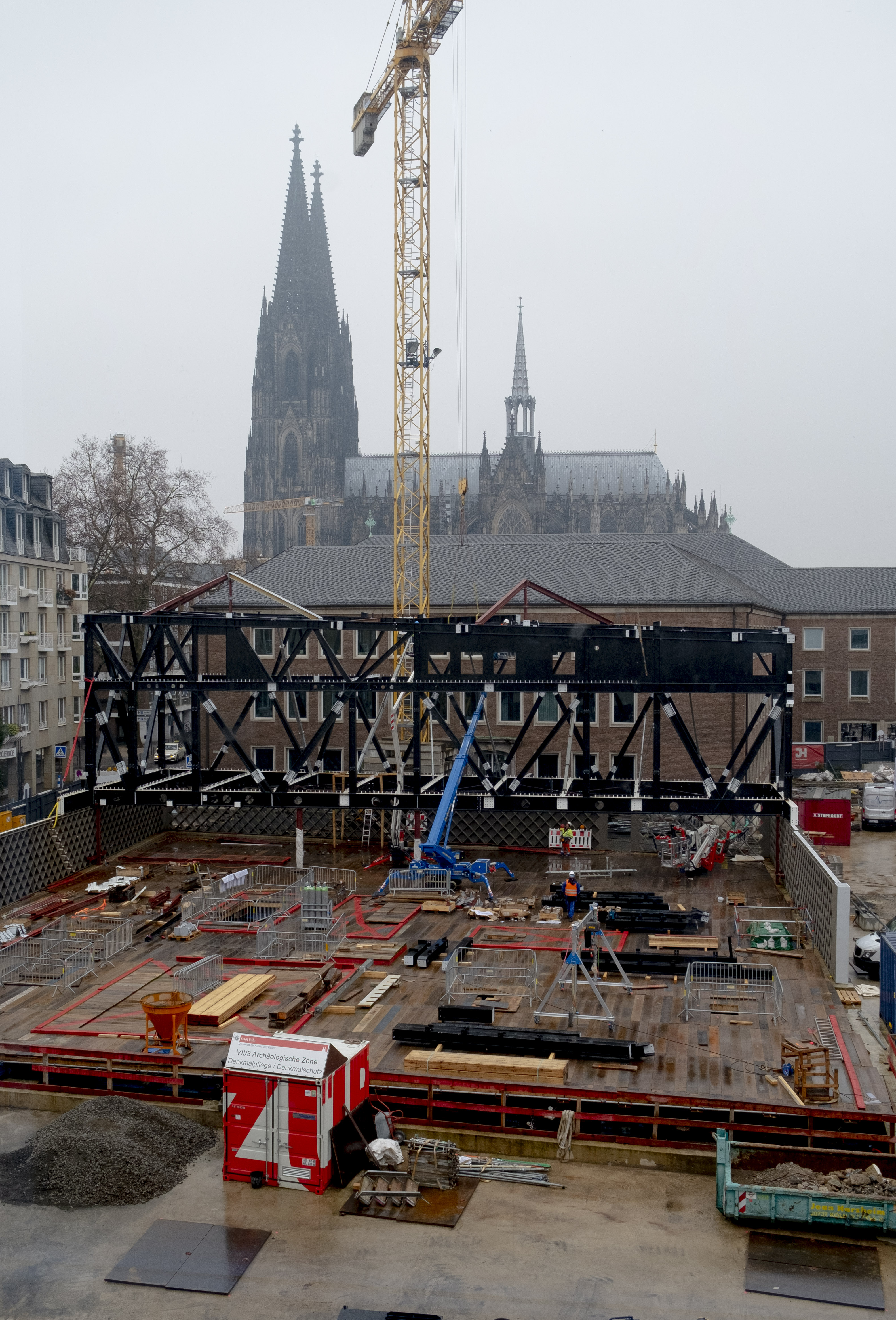 Die MiQua-Baustelle von Süden aus fotografiert. Im Hintergrund ist durch Nebel und vor einem bewölkten Himmel der Kölner Dom zu sehen. Im Zentrum des Bildes liegt die MiQua-Baustelle. Ein hoher gelber Baukran ragt darüber. Die Sicht auf den sogenannten Spanischen Bau wird nun teilweise von einem schwarzen Stahlgerüst verdeckt, das später das Obergeschoss und Dach des Museums halten wird.