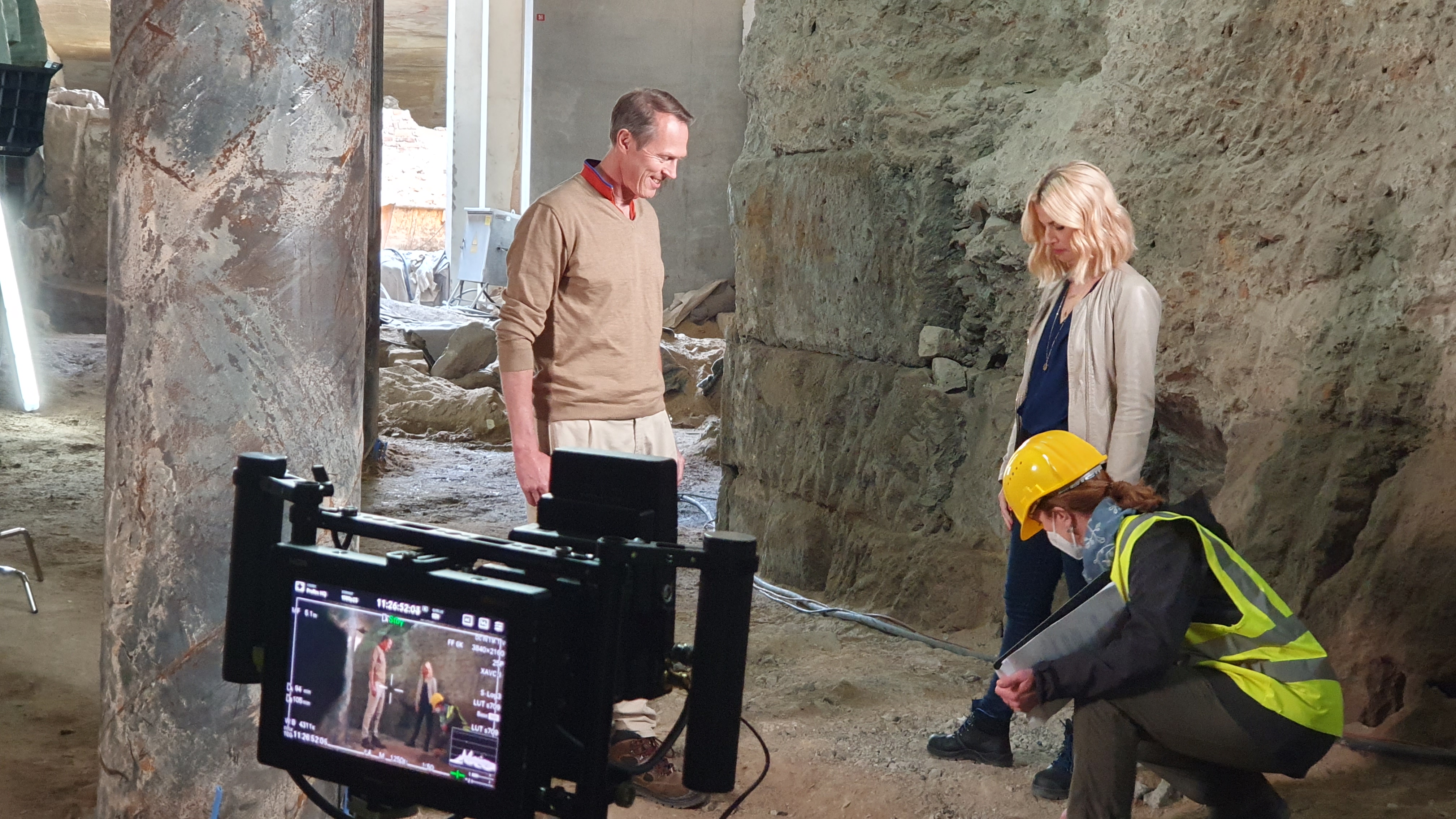 Interviewsituation: Links im Bild Mann mit beige-farbenem Pullover. Frau mit blonden Haaren und beige-farbener Jacke rechts im Bild. Eine Frau mit Bauhelm und Warnweste richtet noch etwas an den Schuhen. Im Vordergrund ist bereits eine Kamera für die Aufnahme ausgerichtet.