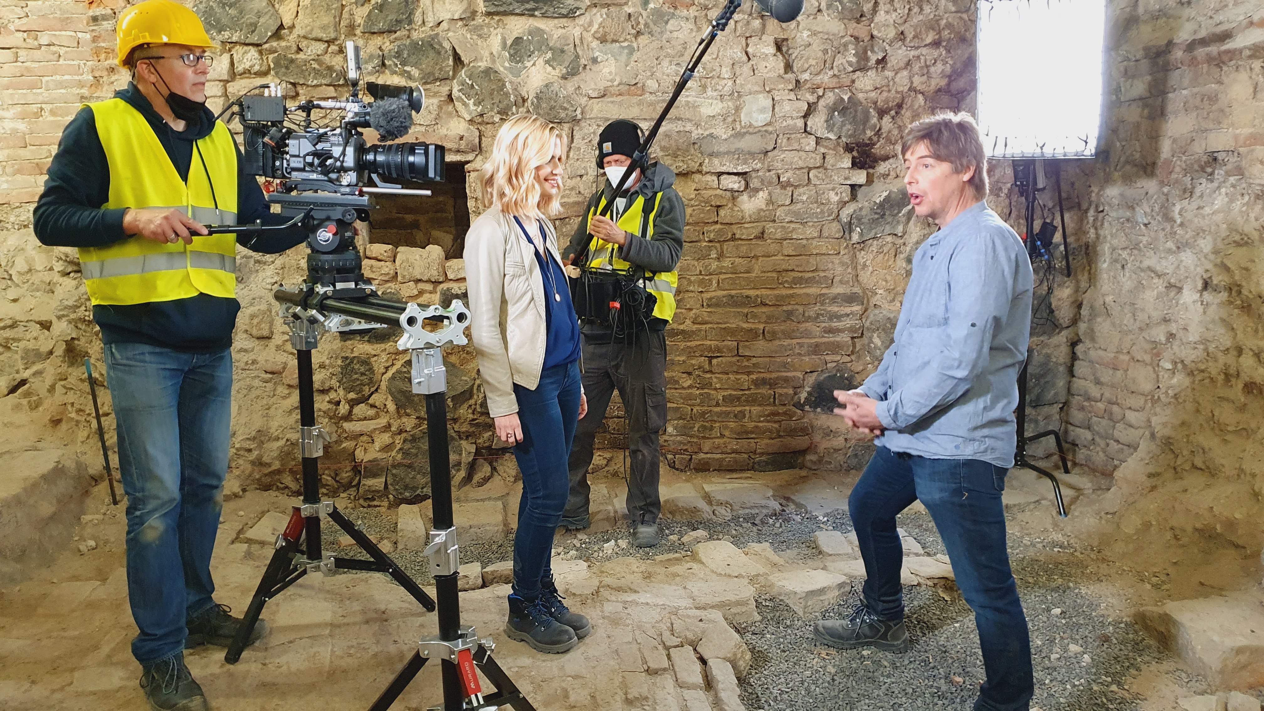 Das Bild zeigt die Situation der Filmaufnahme: Das Kamerateam mit Equipment nimmt das Interview mitten in archäologischen Befunden auf.