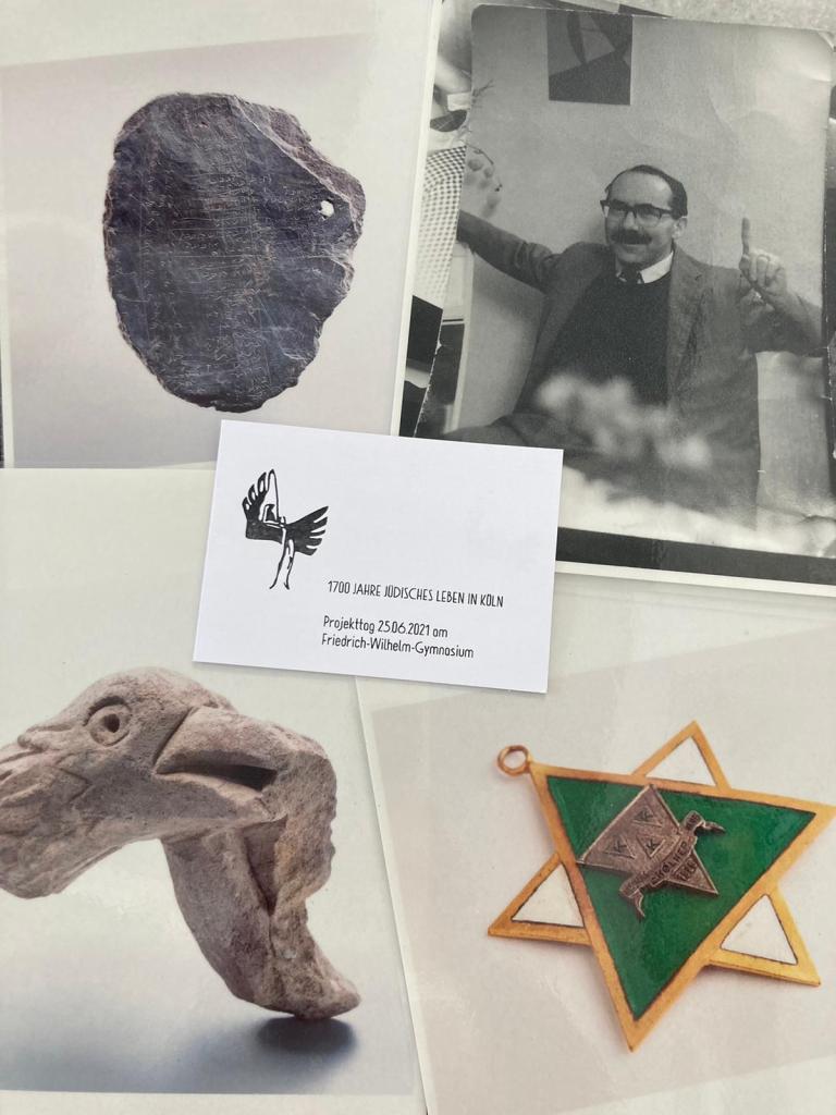 Vier Fotos (mittelalterliche Schiefertafel, Foto eines lachenden Mannes mit Brille und Schnurrbart, steinerner Vogelkopf, Karnevalsorden in Form eines Davidsterns) und eine Einladungskarte mit dem Logo des Friedrich-Wilhelm-Gymnasiums Köln liegen auf einem Tisch.
