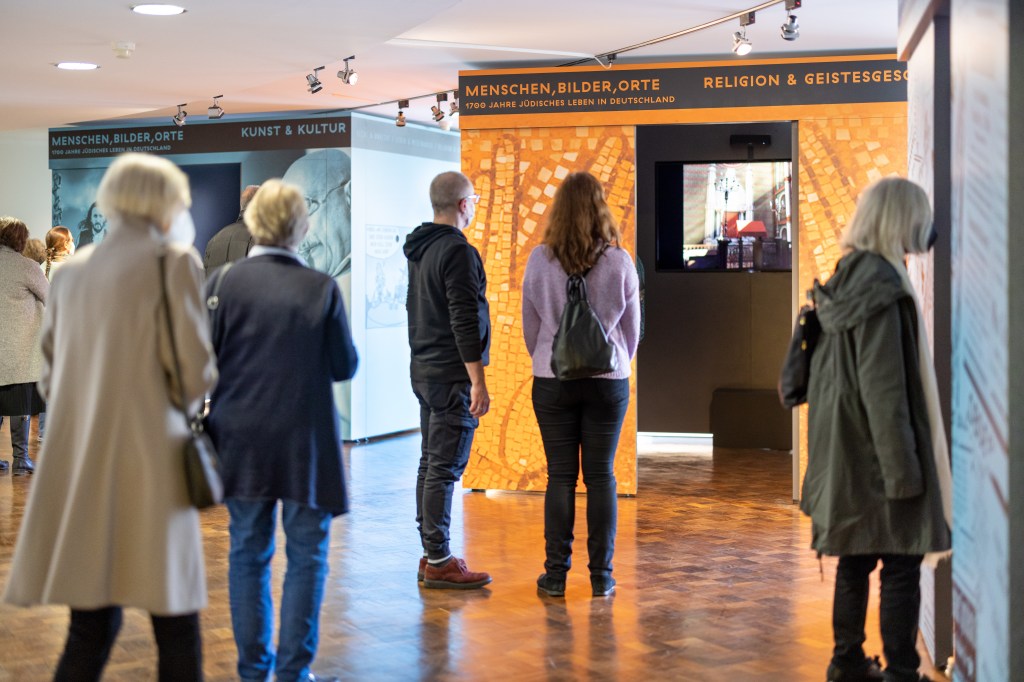 Im Hintergrund die verschieden farbigen Ausstellungskuben. Besucher*innen sehen sich die Inhalte genauer an und füllen den Raum.