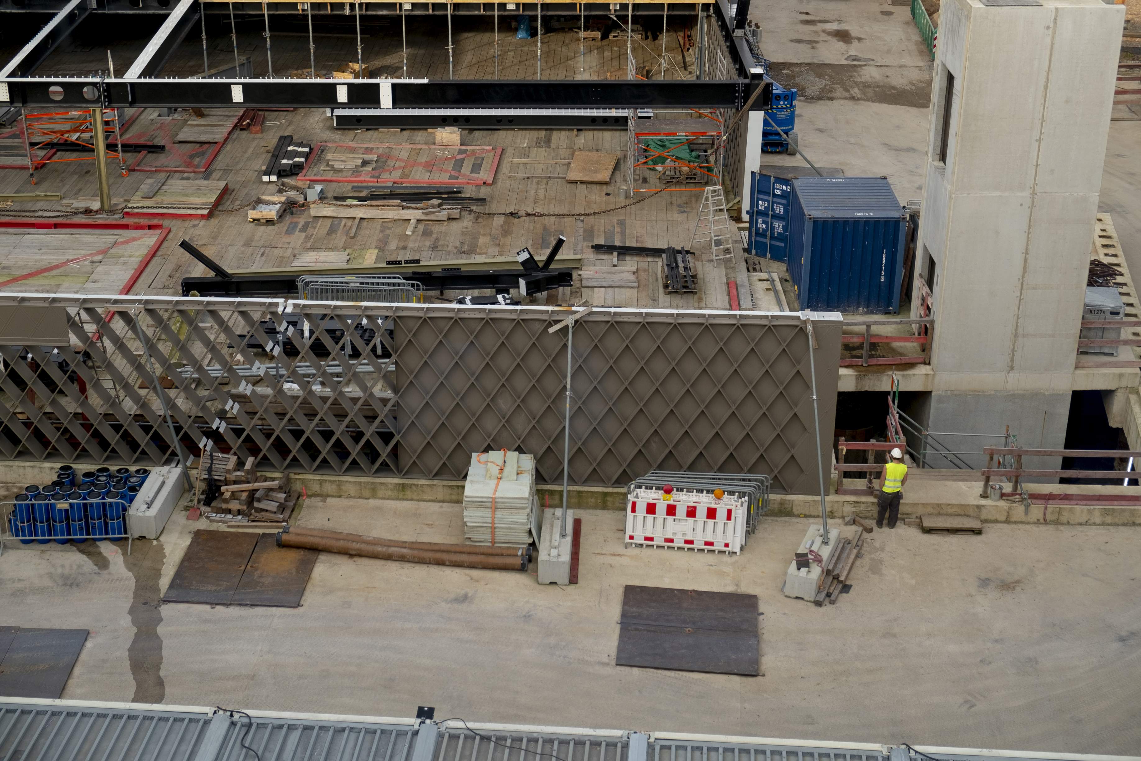 Detailfoto von einem Bereich der Baustelle. Im Zentrum des Bildes steht ein Stück der Stahlkonstruktion. Um das sogenannte Rautentragwerk herum liegen Baumaterialien und Gerätschaften. Ein Bauarbeiter steht neben dem zukünftigen Gebäude.