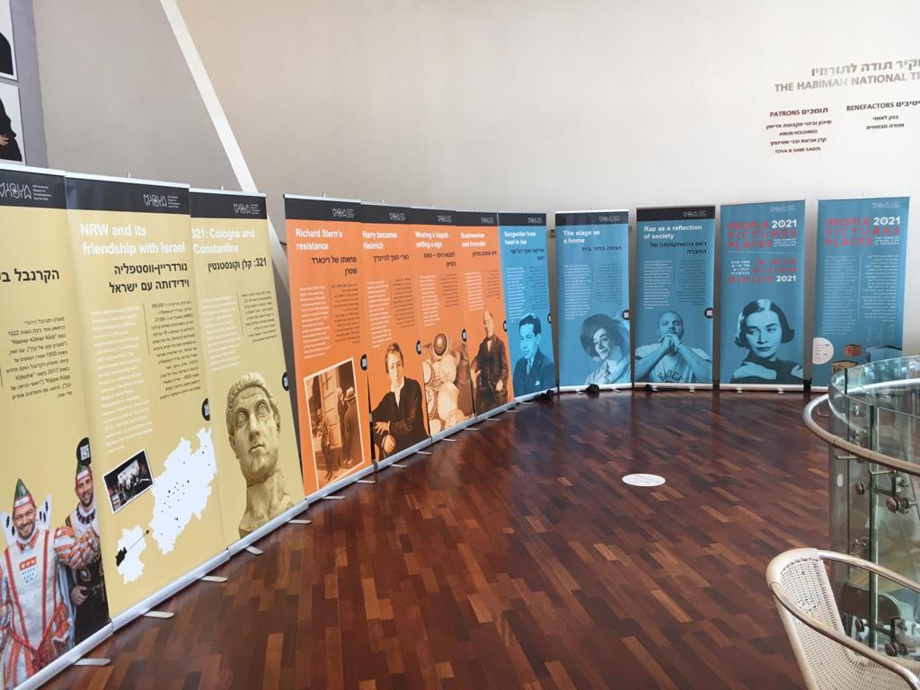 Exhibition space with several roll-ups arranged in a row. The differently coloured posters (yellow, orange, blue) show images of people and texts with biographical backgrounds.