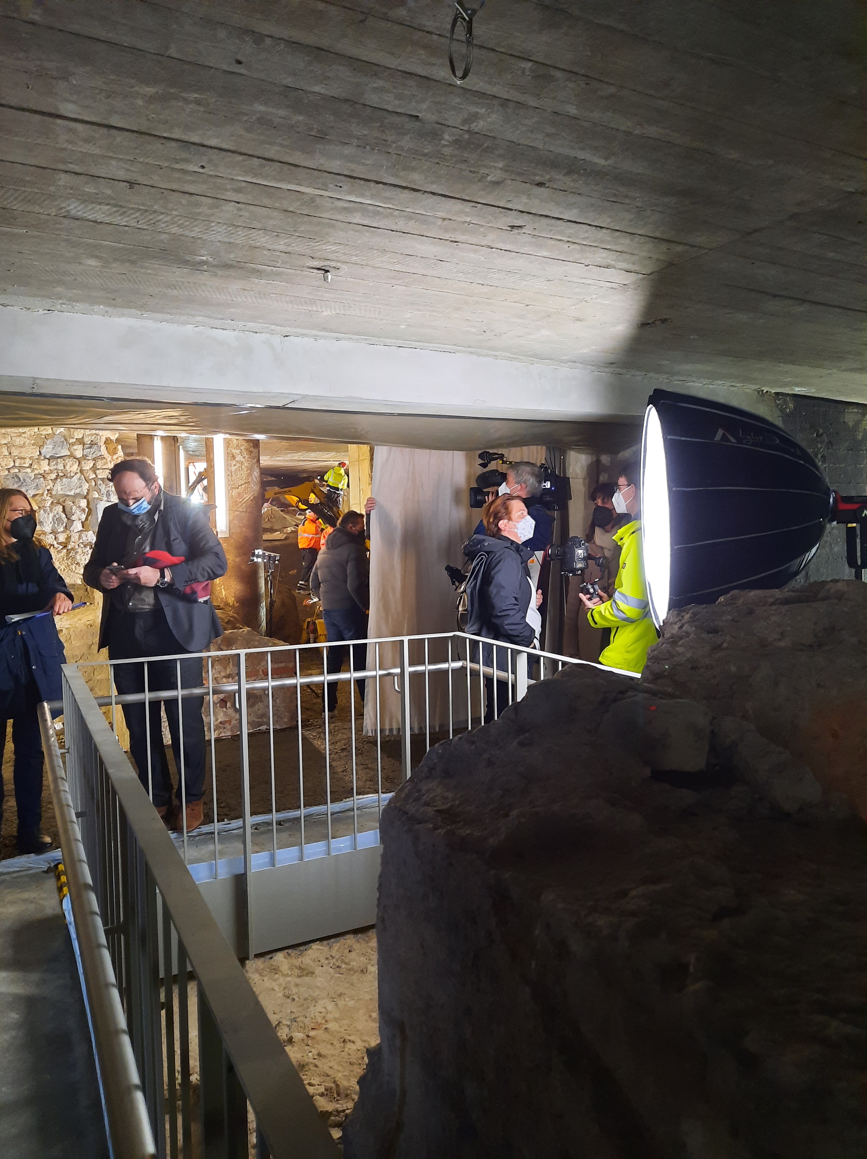 Auf dem Steg über den archäologischen Befunden sammeln sich Fotograf*innen und Kamerateams. Ein großer Lampenschirm sorgt für ausreichende Beleuchtung.