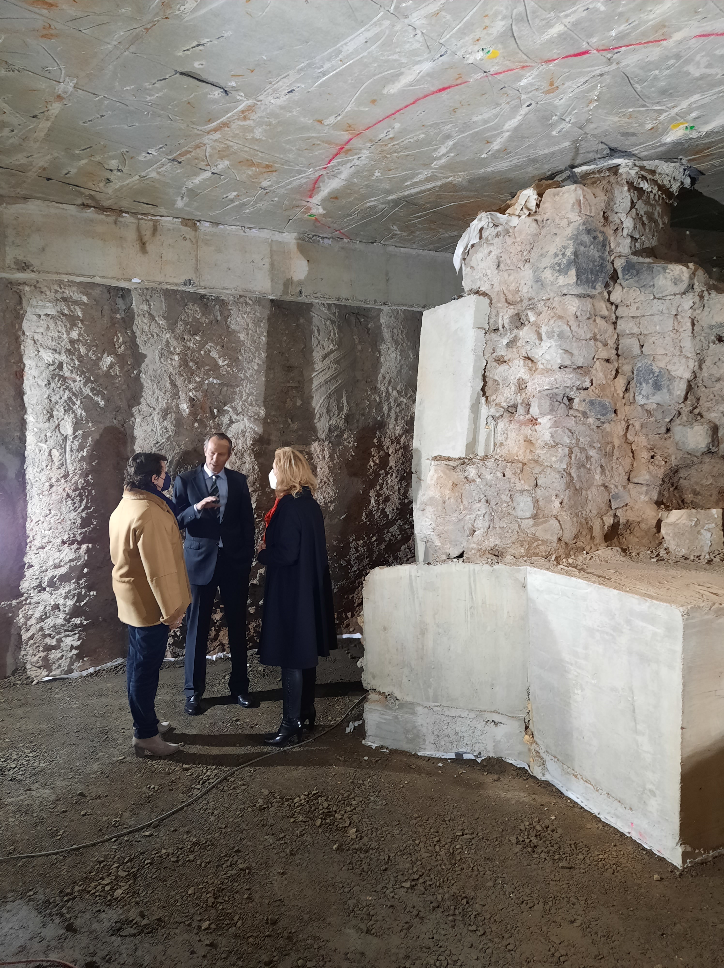 Thomas Otten erläutert Frau Henk-Hollstein und Frau Lubek den Stand der Arbeiten. Sie stehen vor einer Bohrpfahlwand. Rechts im Bild der archäologische Befund.