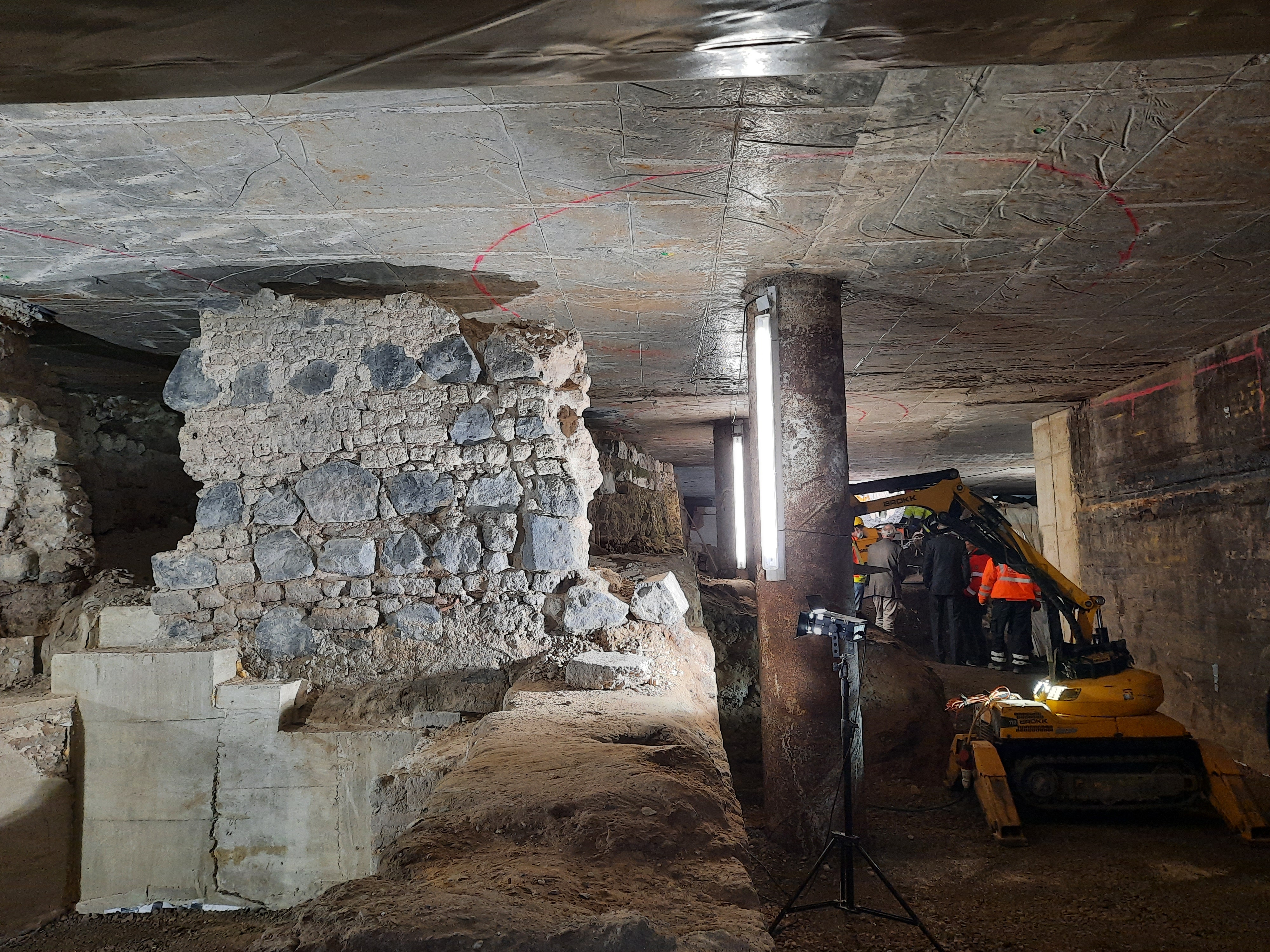 Bauarbeiten in der Befundebene. Film- und Kamerateams machen im Hintergrund Aufnahmen der Arbeiten im Untergrund. In der linken Bildhälfte dominiert archäologischer Befund. Rechts im Bild ein kleiner Bagger.