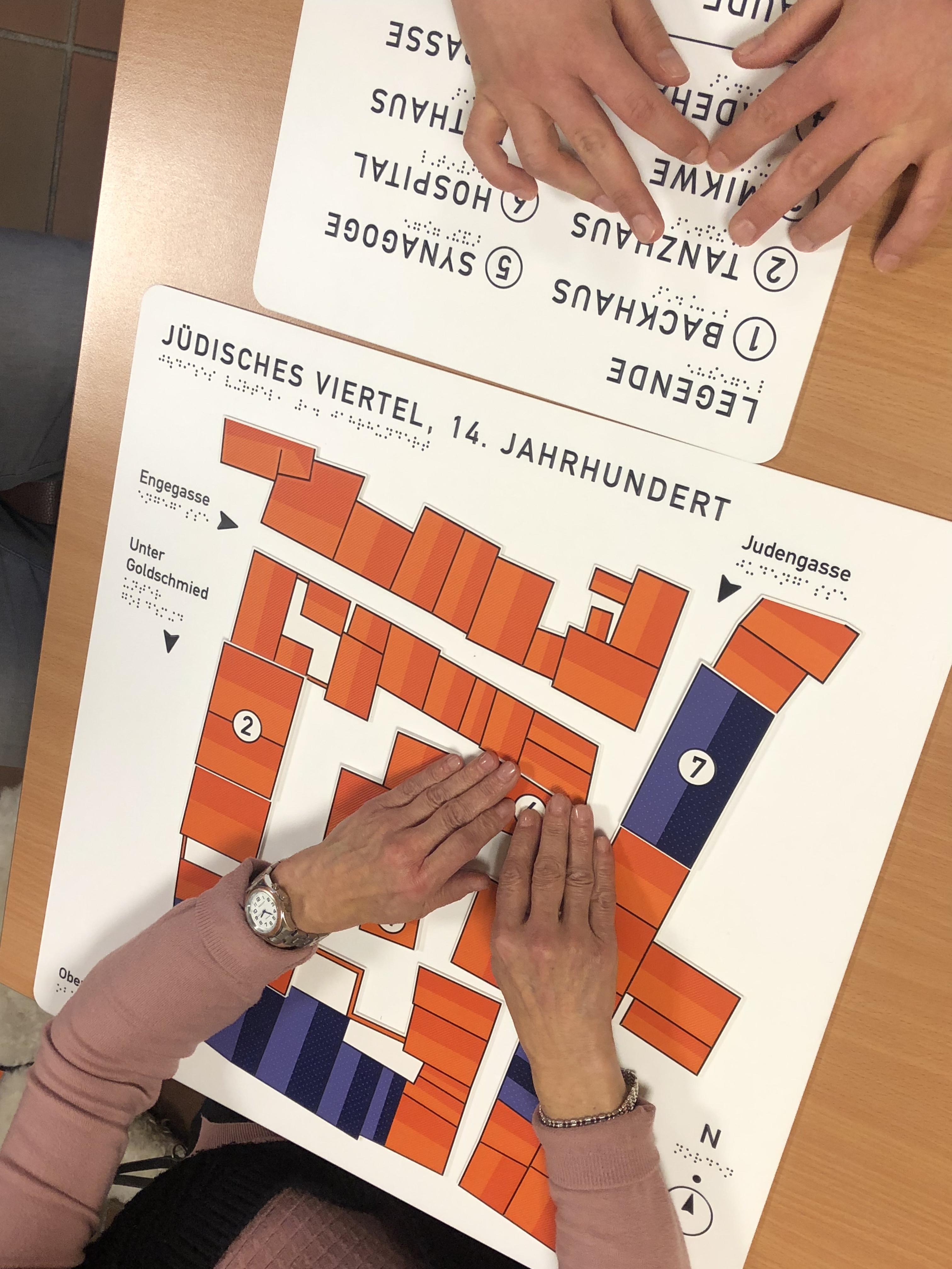 Aufnahme von oben: Auf einem Holztisch liegen zwei Platten mit taktil erfahrbaren Aufschriften und Abbildungen. Zwei sich gegenübersitzende Personen, von denen nur die Hände zu sehen sind ertasten die Modelle.