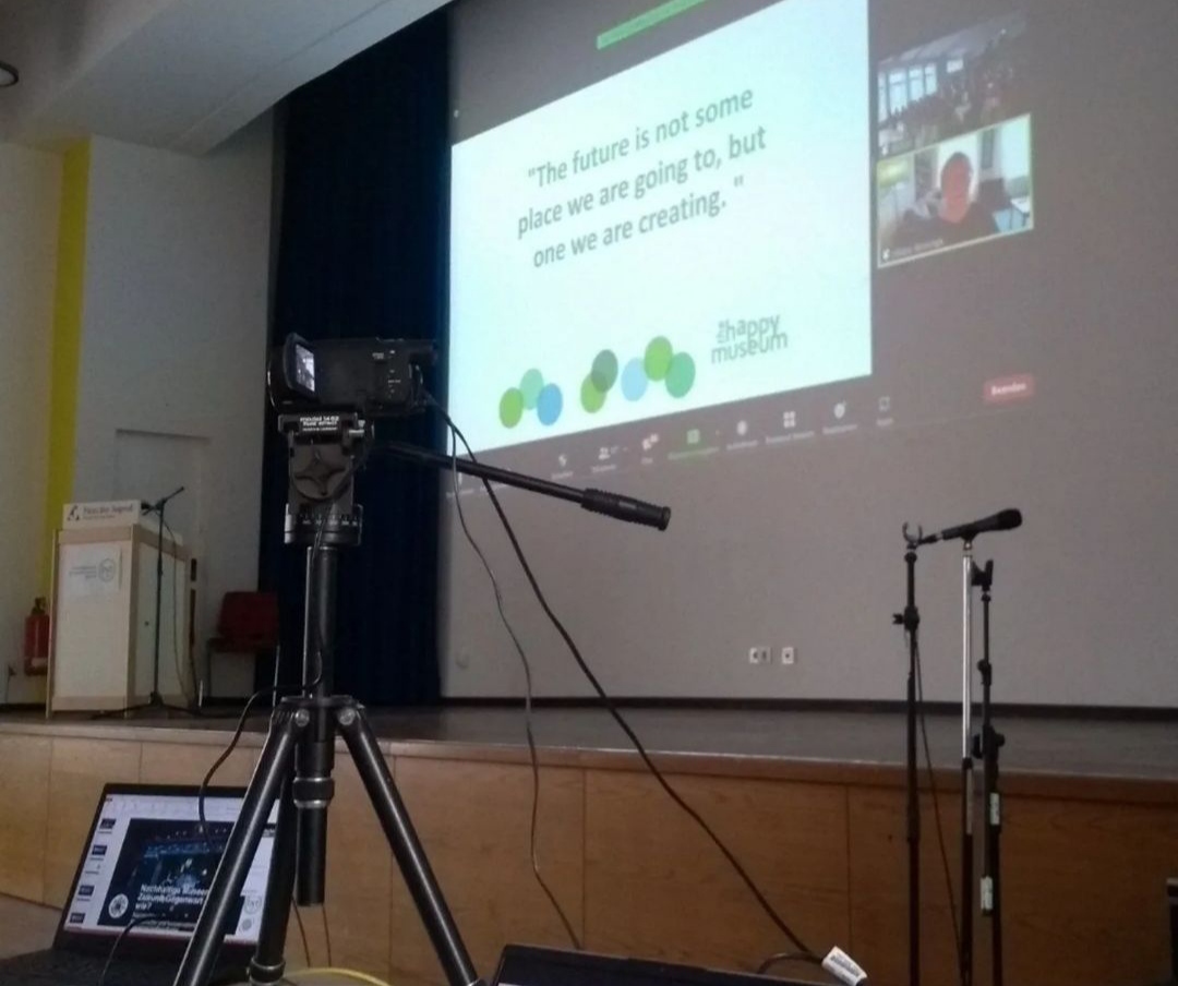 Der Blick auf eine technische Ausrüstung mit Laptop, Kamera und Mikrofon. Im Hintergrund ist auf der Leinwand ein digital zugeschalteter Vortrag erkennbar.