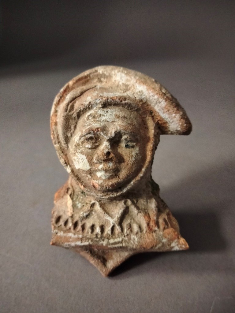 Das Bild zeigt ein Bruchstück einer Ofenkachel in Form einer Männerbüste. Diese trägt eine Kopfbedeckung, die an eine Zipfelmütze erinnert. Die Kachel aus rötlichem Ton zeigt Reste einer grünen Glasur.