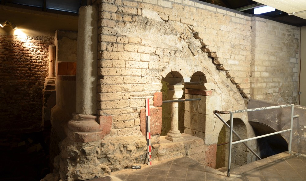 Alte Ziegelmauer mit einer Säule an der linken Wandecke, rechts daneben ein Fenster mit Rundbögen und daneben ein Eisengeländer, das in die Tiefe führt