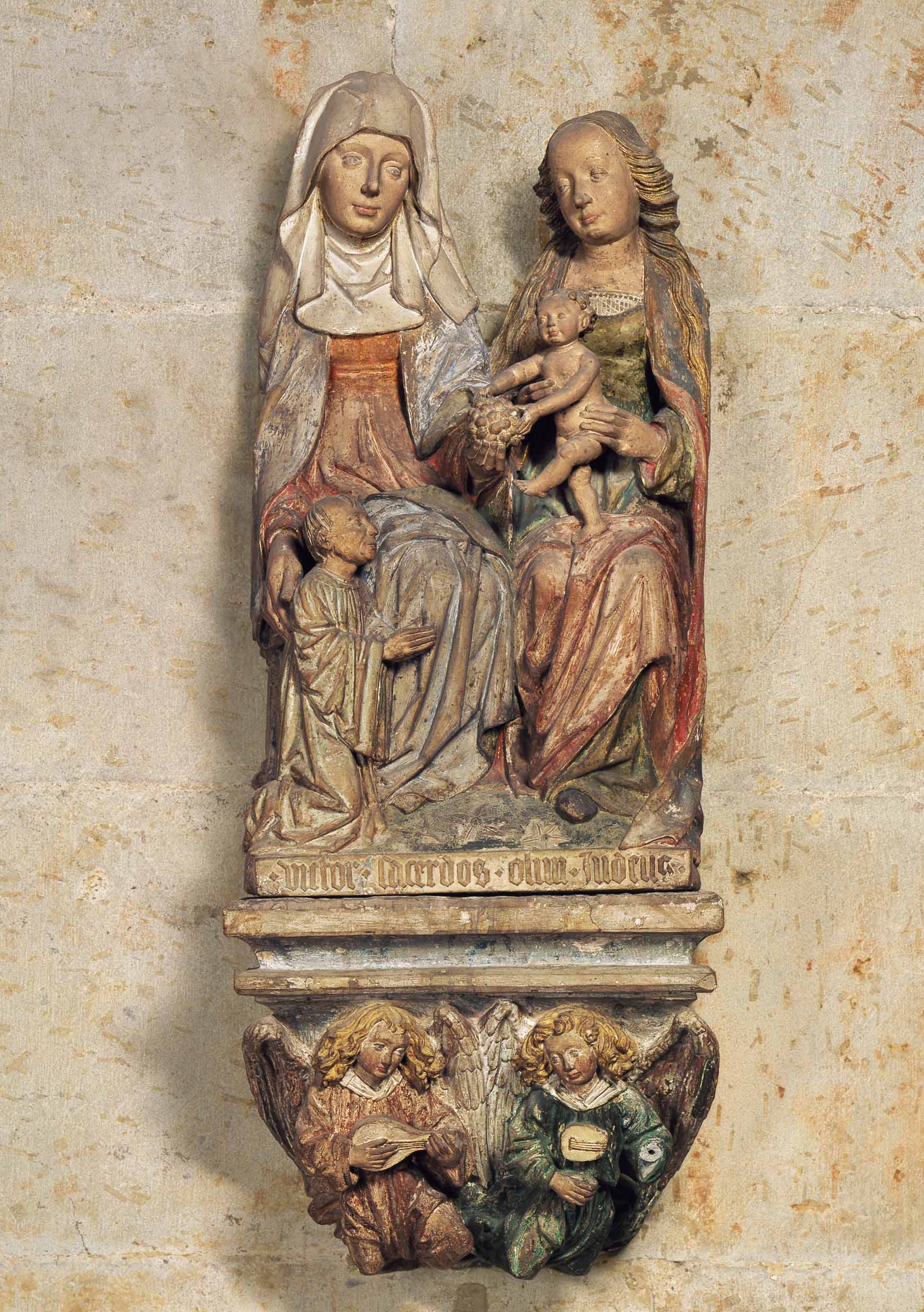 Aufnahme von einer mittelalterlichen Figurengruppe, die aus zwei Frauenfiguren, einem Kleinkind sowie einem knienden Mann zu ihren Füßen und zwei Engelsgestalten am unteren Ende besteht. Ein Schriftband weißt auf den Stifter hin. Sie hängt in einer Seitenkapelle des Kölner Doms.