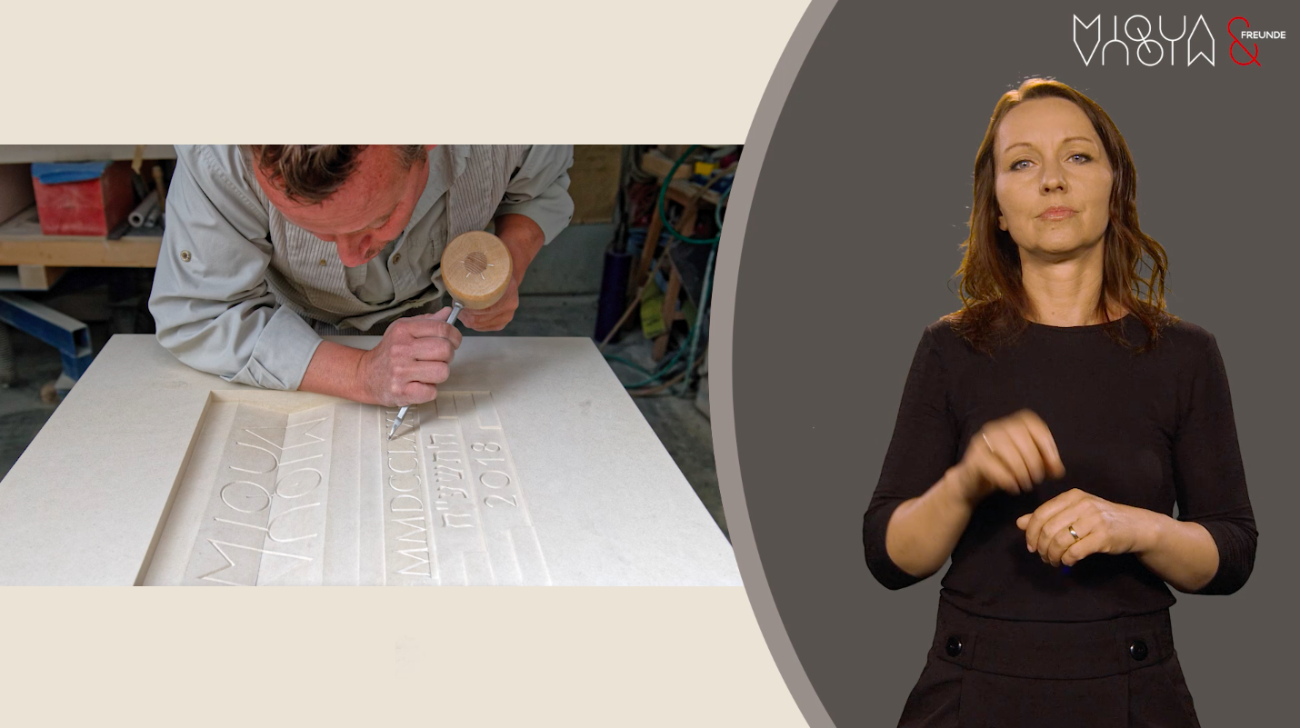 Bildschirmaufnahme eines Videobeitrags. Links im Bild ein Foto, das zeigt, wie der Grundstein des MiQua mit einem Meißel bearbeitet wird. Rechts im Bild steht eine Frau in schwarzer Kleidung und mit schulterlangen braunen Haaren. Mit ihren Händen dolmetscht sie das gesprochene Wort in Deutsche Gebärdensprache.