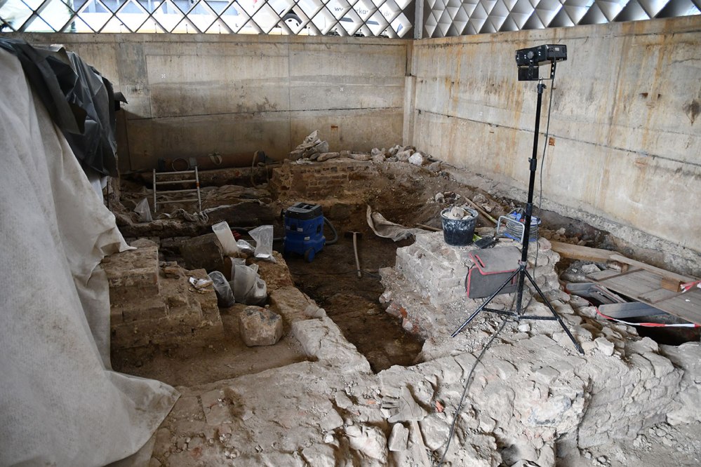 Vor glatten grauen Betonwänden sieht man die Fundamentreste antiker Gebäude, links sind Teile der Gemäuer mit Plastikfolie abgedeckt. Überall liegen die Werkzeuge der Archäologen herum, darunter Eimer, Kabeltrommeln, eine Schaufel und rechts steht ein Scheinwerfer auf einem Stativ