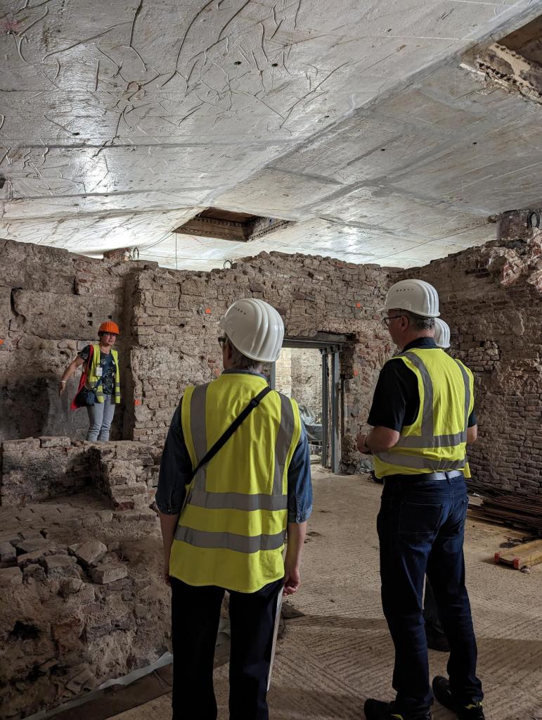 In einem Raum mit alten Steinmauern stehen im Vordergrund drei Personen mit gelben Warnwesten und weißen Bauhelmen. Im Hintergrund steht eine vierte Person mit einem orangenen Bauhelm und erklärt etwas