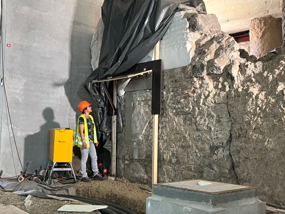 Eine Frau mit gelber Warnweste und einem orangenen Bauhelm steht vor einer Betonmauer, neben ihr ein gelber Metallkasten. Rechts von Ihr in einer alten Steinwand befindet sich eine Inschrift, deren Abdeckung aus Holz geöffnet ist