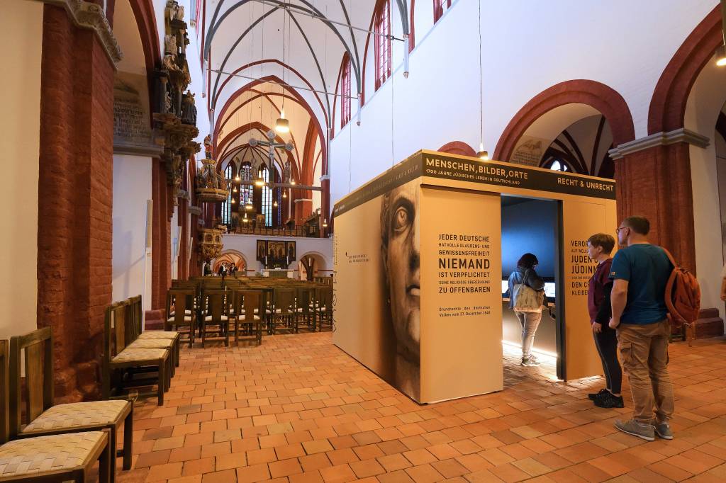 Eine Frau steht in einem Ausstellungskubus im Dom Sankt Peter und Paul. Zwei Männer stehen davor und schauen durch die Tür ins Innere des Kubus