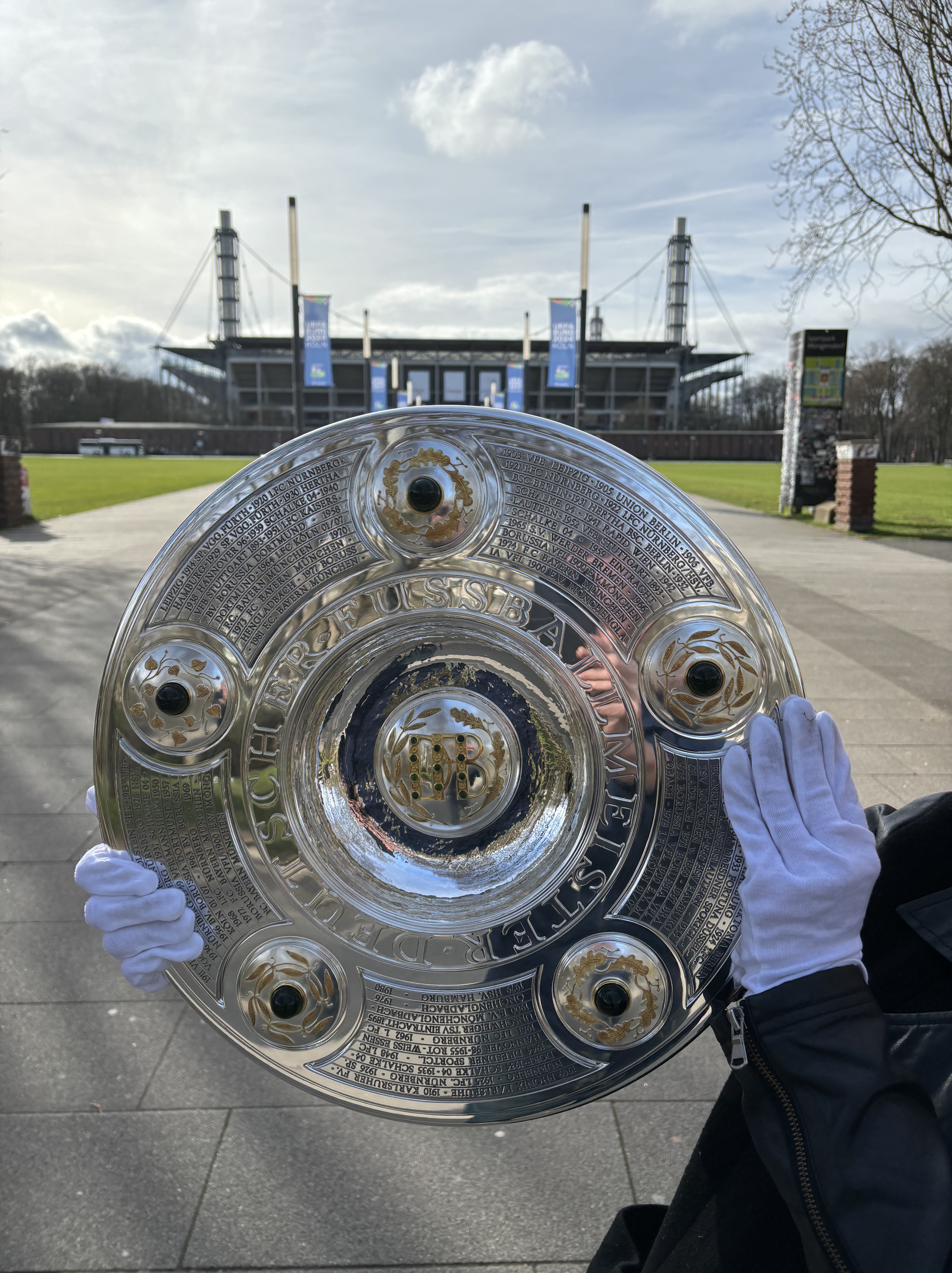 Aufnahme des Stadions in Köln. Davor hält eine Person die Meisterschale, ein silbernes Objekt, nach oben.
