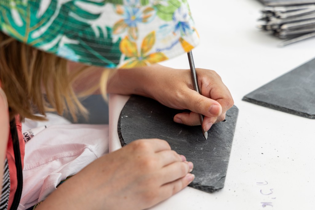 Ein junges Kind ritzt und kratzt auf einer Schiefertafel. Es trägt einen bunten Hut.