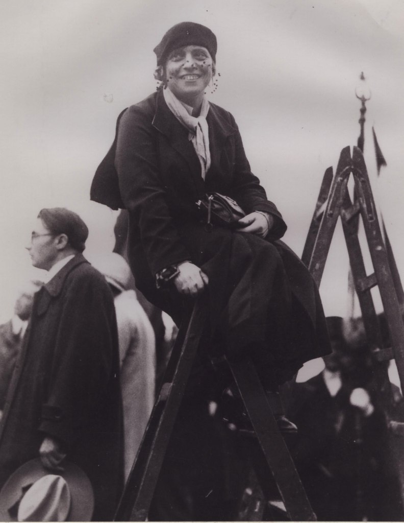 Eine Frau sitzt lächelnd auf einer hohen Konstruktion, möglicherweise einer Leiter oder einem Gerüst. Sie trägt einen dunklen Mantel, einen Schal und eine Baskenmütze. Im Hintergrund stehen weitere Personen, von denen einige in Mänteln und Hüten gekleidet sind. Das Bild hat eine historische Atmosphäre und ist in Schwarz-Weiß gehalten.