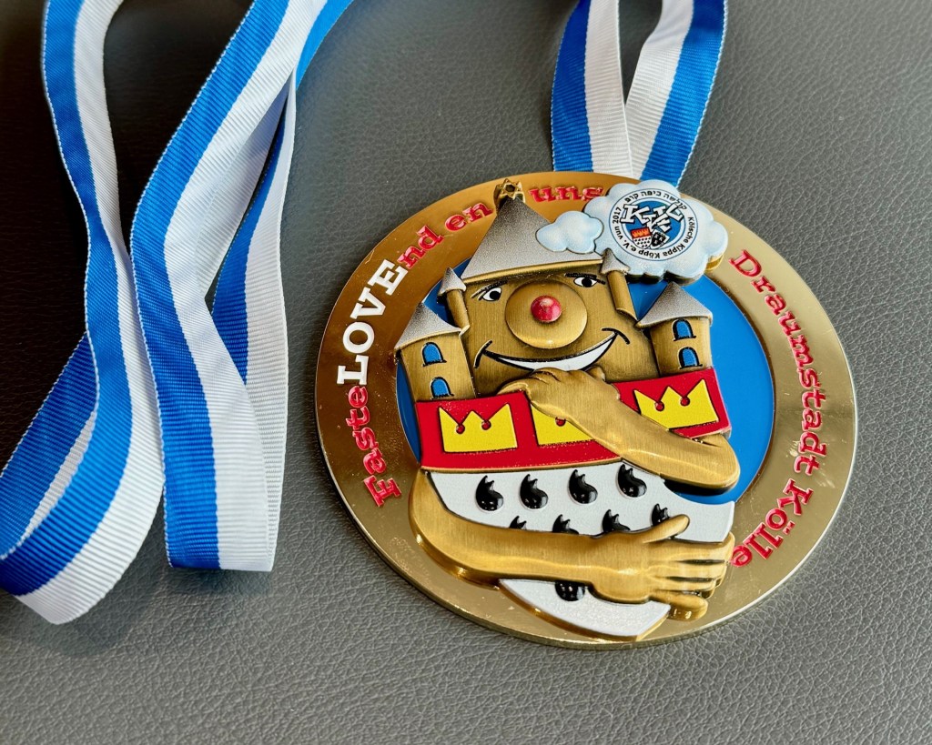 Ein farbenfroher Karnevalsorden mit blau-weißem Band liegt auf einer grauen Oberfläche. Das Motiv zeigt eine fröhliche, schlossähnliche Figur, die das Kölner Stadtwappen umarmt. Die Aufschrift „FasteLOVEnd en uns Draumstadt Kölle“ verbindet den Kölner Karneval mit einer Botschaft der Gemeinschaft und Freude. Ein kleiner Pin mit hebräischen Zeichen und einem Karnevalsemblem ergänzt das Design und verweist auf die kulturelle Vielfalt des Kölner Karnevals.