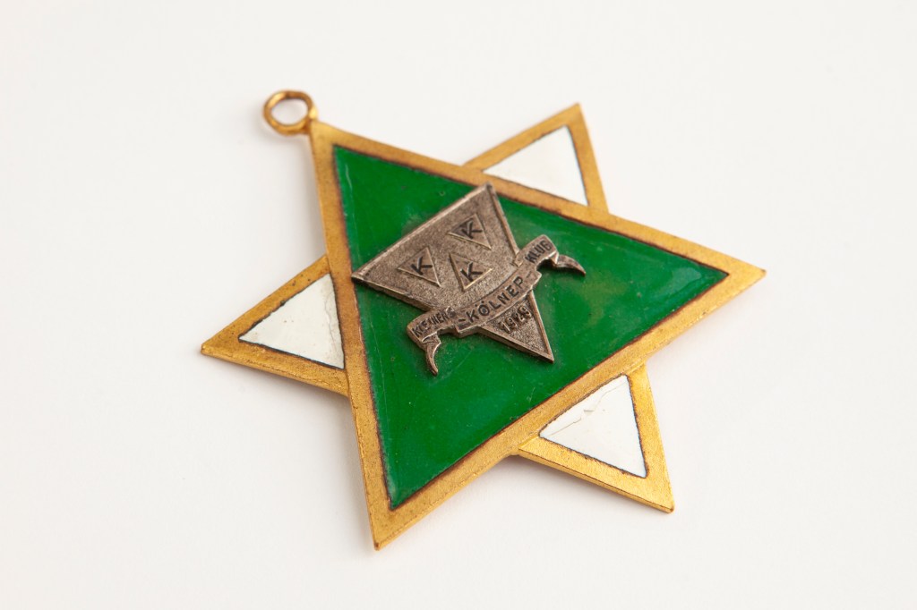 Ein historischer Karnevalsorden in Form eines Davidsterns mit goldenen, grünen und weißen Elementen. In der Mitte befindet sich ein Metallemblem mit der Aufschrift „Kleiner Kölner Klub 1929“. Dieses Abzeichen verbindet Karnevalstradition mit jüdischer Symbolik und verweist auf die kulturelle Vielfalt und Geschichte des Kölner Karnevals.