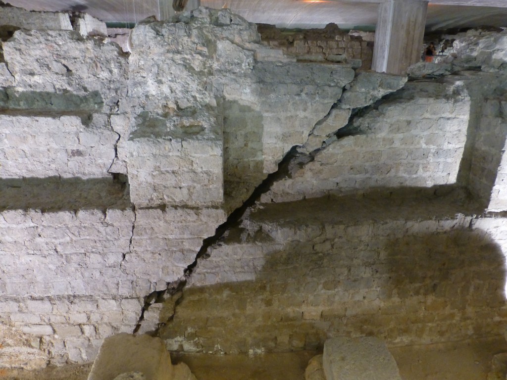 Die Abbildung zeigt eine historische Mauer mit einer markanten diagonalen Rissbildung. Die aus Ziegelsteinen und Mörtel errichtete Struktur gehört zu einer archäologischen Stätte und weist sichtbare Schäden auf, die auf Setzungen oder andere statische Einwirkungen hindeuten. Im oberen Bereich sind moderne Stützen zu erkennen, die möglicherweise zur Sicherung der historischen Bausubstanz dienen. Licht- und Schatteneffekte betonen die Struktur der Mauer und die Tiefe des Risses.