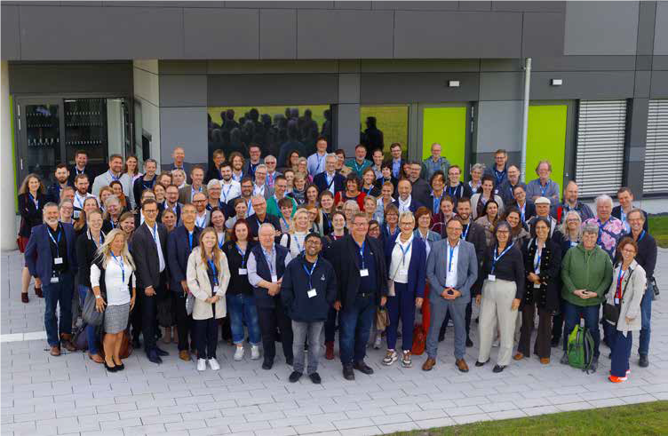 Ungefähr 100 Menschen stehen vor einem modernen Gebäude und blicken hoch in die Kamera.