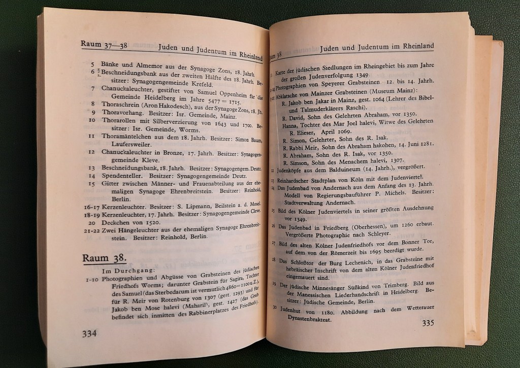 „Doppelseite aus dem Ausstellungskatalog der Jahrtausend-Ausstellung der Rheinlande, Köln 1925. Gezeigt wird das Verzeichnis der Exponate in den Räumen 37 und 38, die dem Thema ‚Juden und Judentum im Rheinland‘ gewidmet waren. Die Auflistung umfasst religiöse Kultgegenstände wie Chanukkaleuchter, Thoravorhänge, Beschneidungsbänke und Grabsteinabbildungen, die teils aus bedeutenden Synagogengemeinden der Region stammen. Die Vielfalt der Objekte dokumentiert eindrucksvoll das reiche jüdische Erbe im Rheinland.